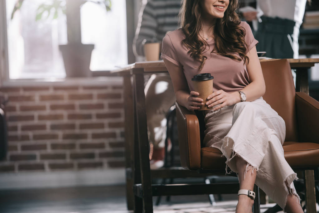 vista recortada de la sonriente mujer de negocios sentada en sillón con café para ir cerca de colegas
 - Foto, Imagen