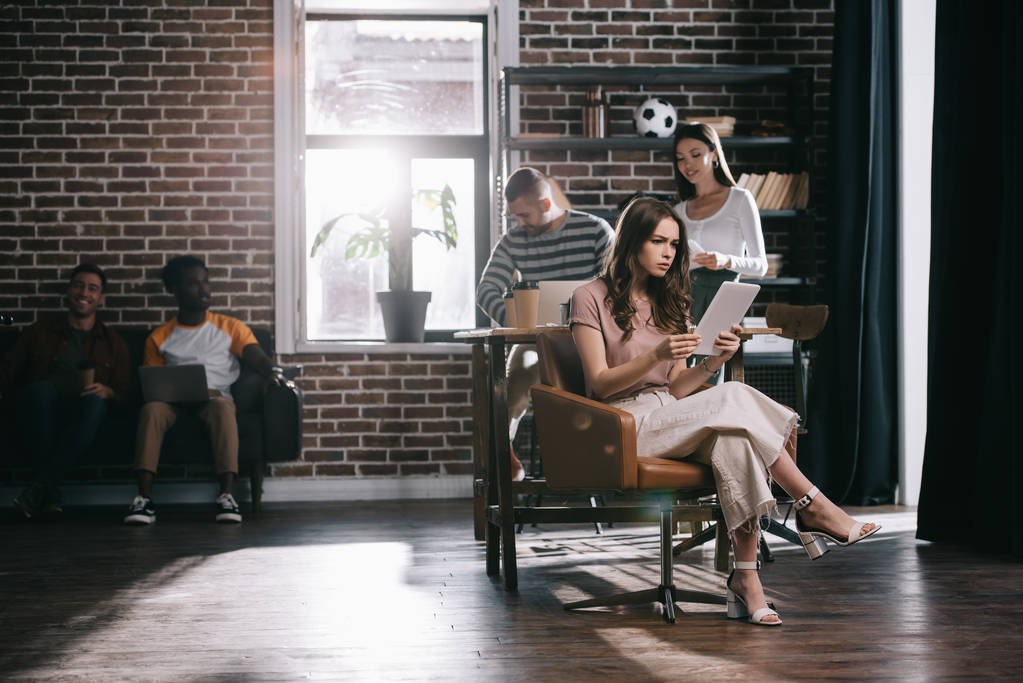 сконцентрированная деловая женщина, сидящая в кресле и использующая цифровой планшет рядом с молодыми мультикультурными коллегами
 - Фото, изображение
