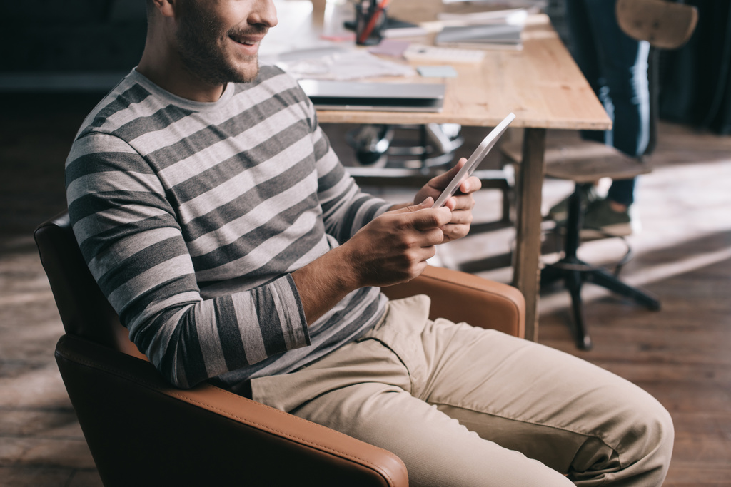 обрезанный вид улыбающегося бизнесмена с помощью цифрового планшета во время сидения в кресле
 - Фото, изображение