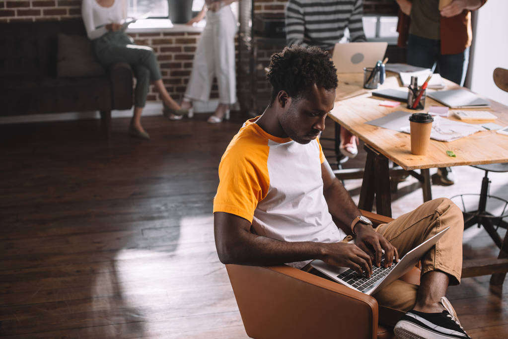 アフリカ系アメリカ人のビジネスマンがノートパソコンを使いコーヒーを飲みながら机の近くのアームチェアに座り - 写真・画像