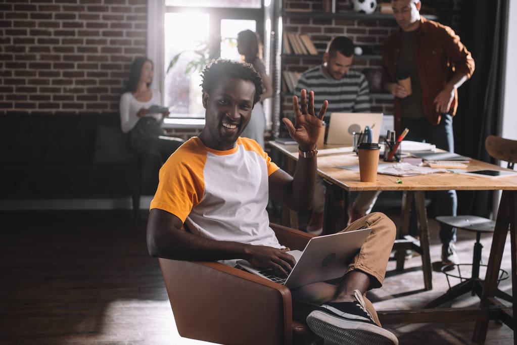 χαμογελαστός Αφροαμερικάνος επιχειρηματίας που χρησιμοποιεί φορητό υπολογιστή ενώ κάθεται στην πολυθρόνα και χαιρετάει δίπλα σε νέους πολυπολιτισμικούς συναδέλφους - Φωτογραφία, εικόνα