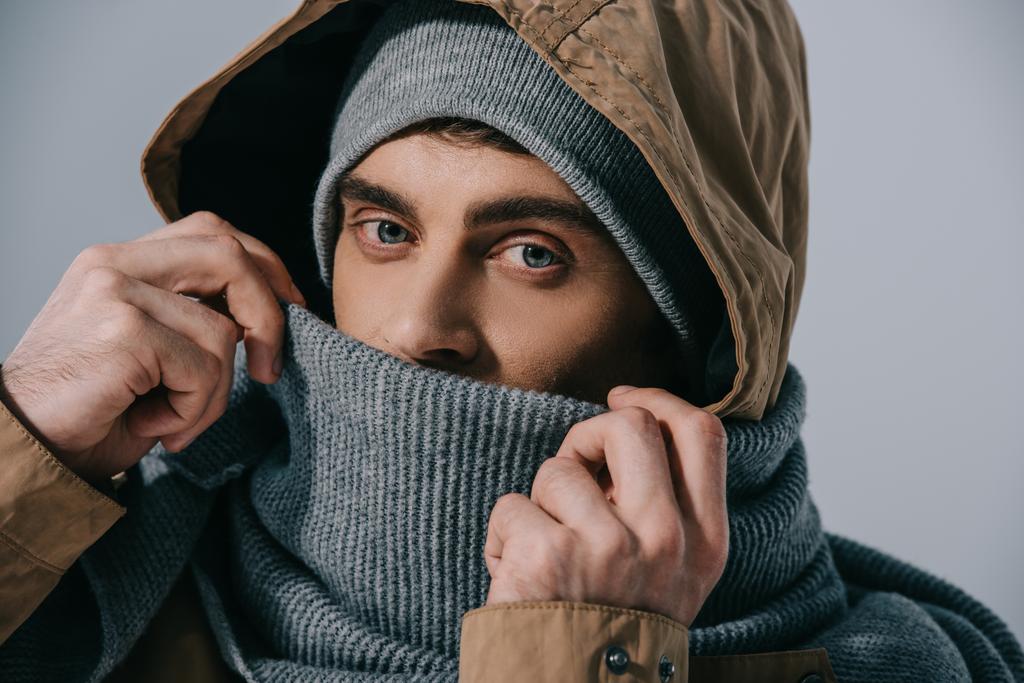 pohledný mladý muž pózující v klobouku, šále a kapuci, izolovaný na šedi - Fotografie, Obrázek
