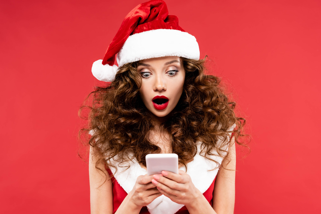 shocked woman in santa costume using smartphone, isolated on red - Photo, Image