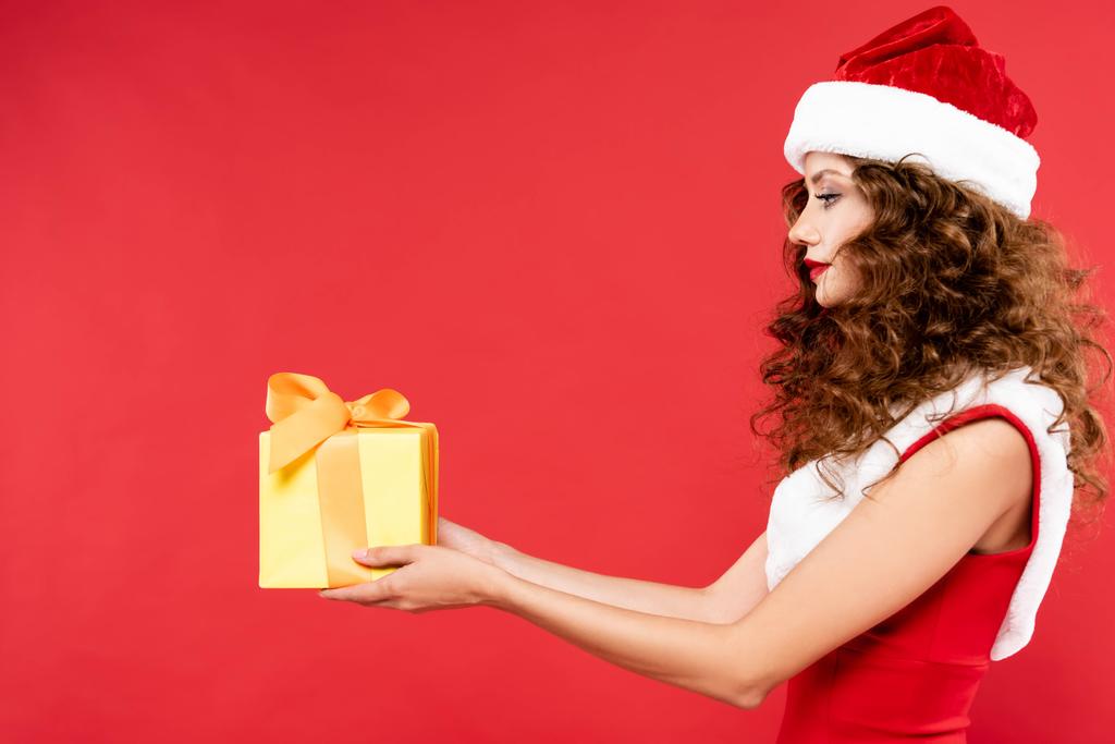 кудрявая девушка в костюме Санты, держит рождественский подарок, изолированный на красный
 - Фото, изображение