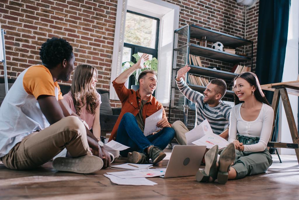 gelukkige zakenmensen geven high five terwijl zitten op de vloer in de buurt van jonge multiculturele collega 's - Foto, afbeelding