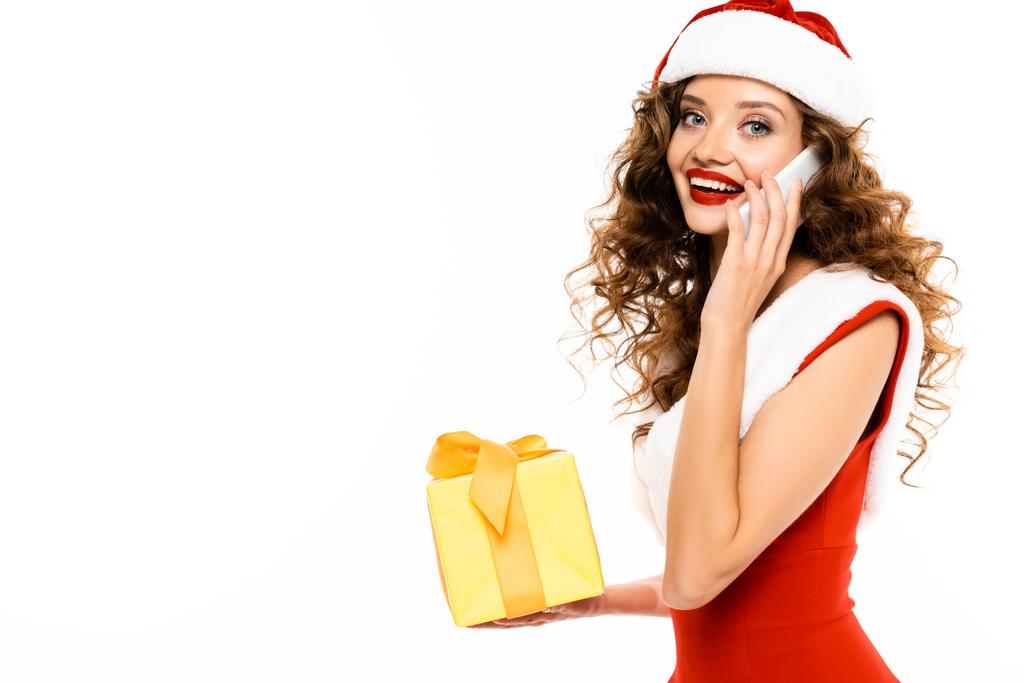atraktivní dívka v kostýmu Santa drží vánoční dárek a mluví na smartphone, izolované na bílém - Fotografie, Obrázek