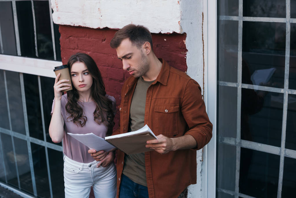 tired businesswoman holding coffee to go near head while standing near thoughtful colleague with documents - Photo, Image