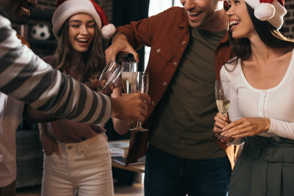 bijgesneden beeld van zakenman gieten champagne in glazen van gelukkige collega 's in santa hoeden - Foto, afbeelding