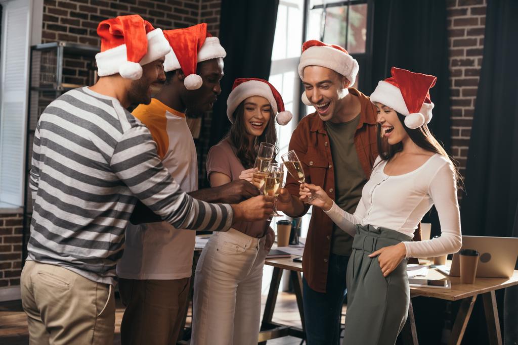 happy multicultural businesspeople in santa hats clinking champagne glasses in office - Photo, Image