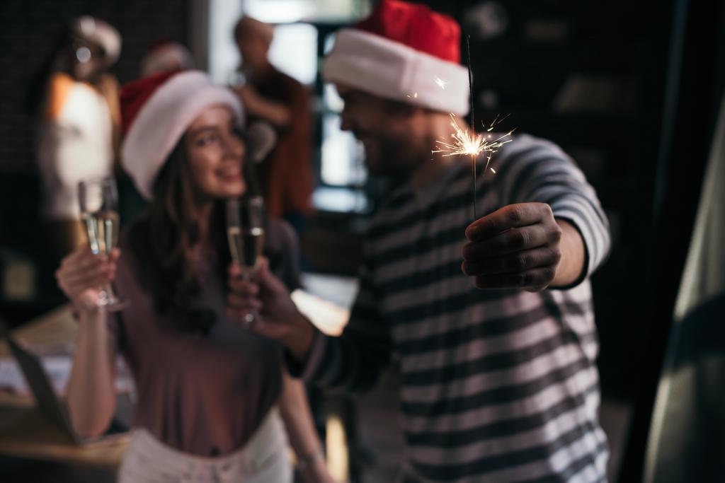 selective focus of happy businessman and businesswoman in santa hats looking at each other while holding champagne glasses - Photo, Image