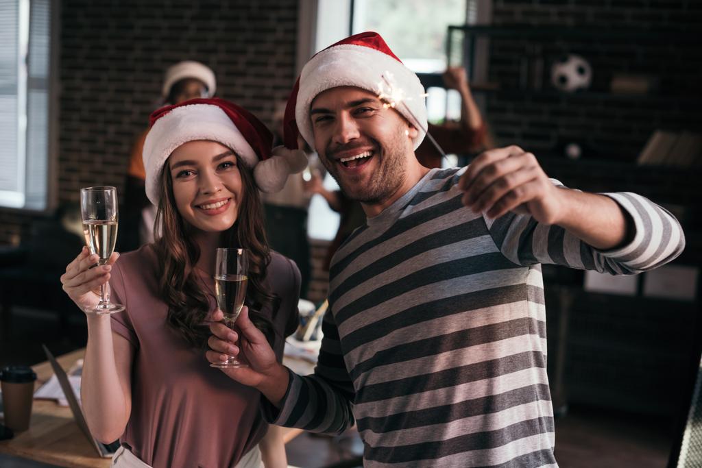homme d'affaires heureux et femme d'affaires dans chapeaux Santa regardant la caméra tout en tenant des verres de champagne
 - Photo, image