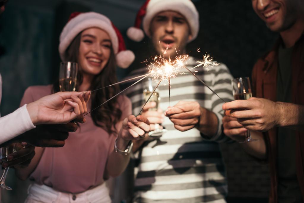 fröhliche, junge Geschäftsleute in Weihnachtsmannhüten zünden Wunderkerzen an, während sie Champagnergläser in der Hand halten - Foto, Bild