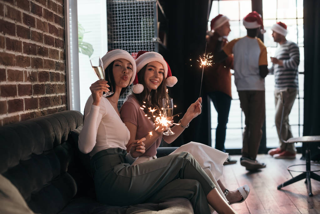 young, happy businesswoman in santa hats smiling at camera while sitting on sofa with sparklers and champagne glasses - Photo, Image