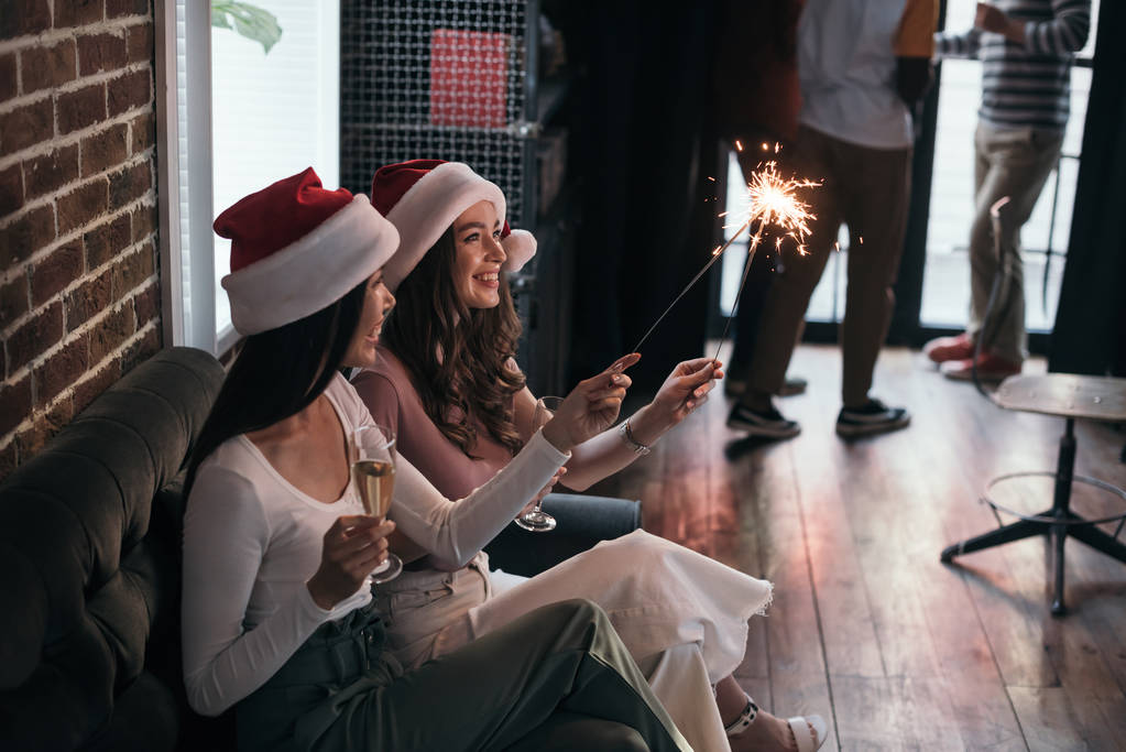 junge, fröhliche Geschäftsfrau in Weihnachtsmannmützen auf Sofa mit Wunderkerzen und Sektgläsern - Foto, Bild