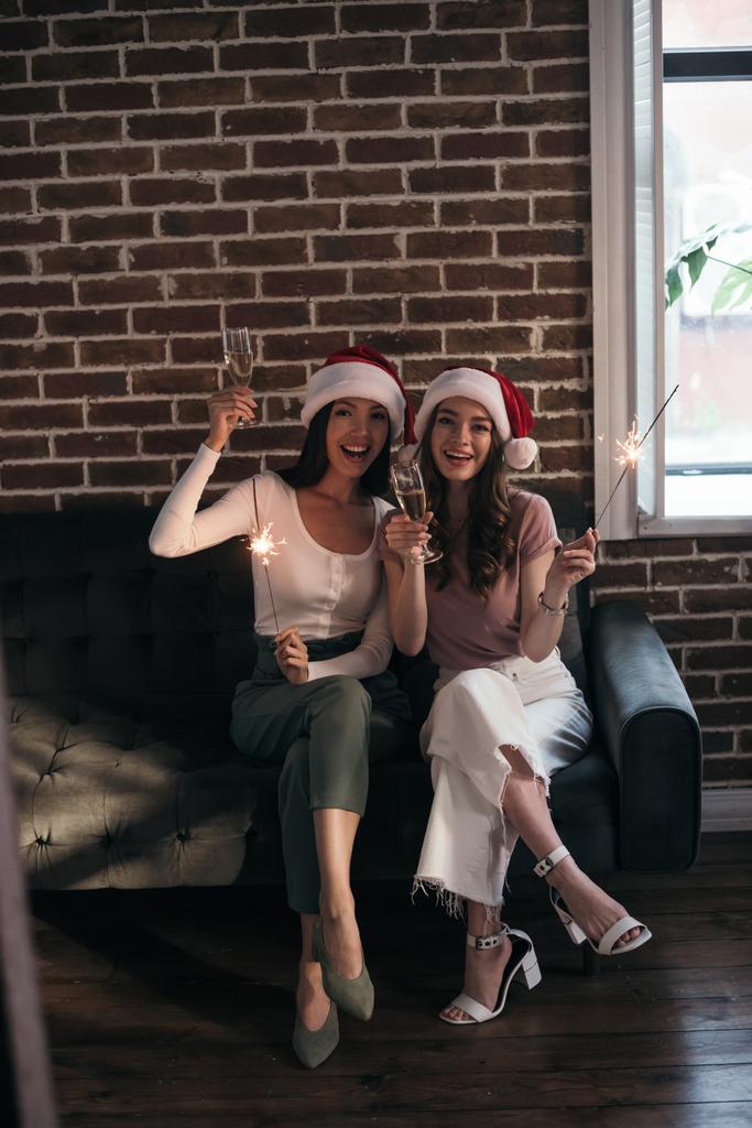 two young, happy businesswoman in santa hats smiling at camera while sitting on sofa with sparklers and champagne glasses - Photo, Image