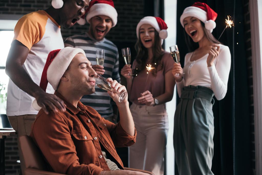 mladý podnikatel pije šampaňské, zatímco stojí poblíž veselých multikulturních kolegů v Santa Klobouky - Fotografie, Obrázek