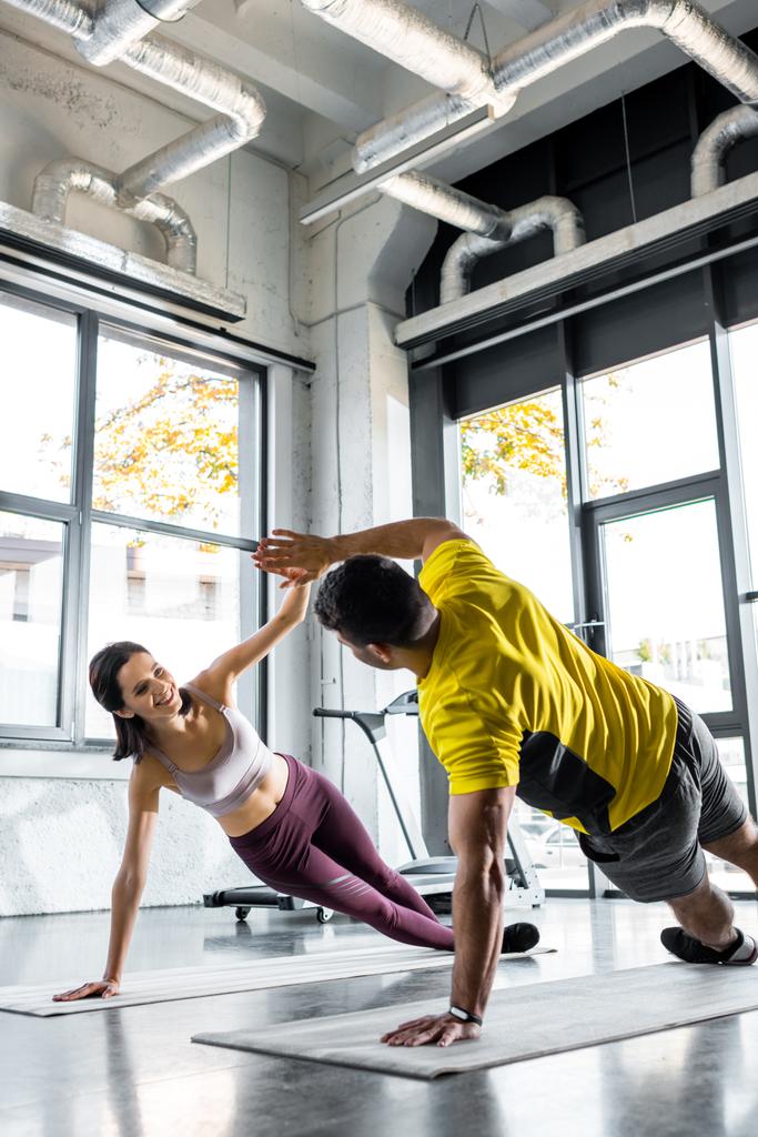 sportman en lachende sportvrouw die plank doet en klapt op fitnessmatten in sportcentrum - Foto, afbeelding
