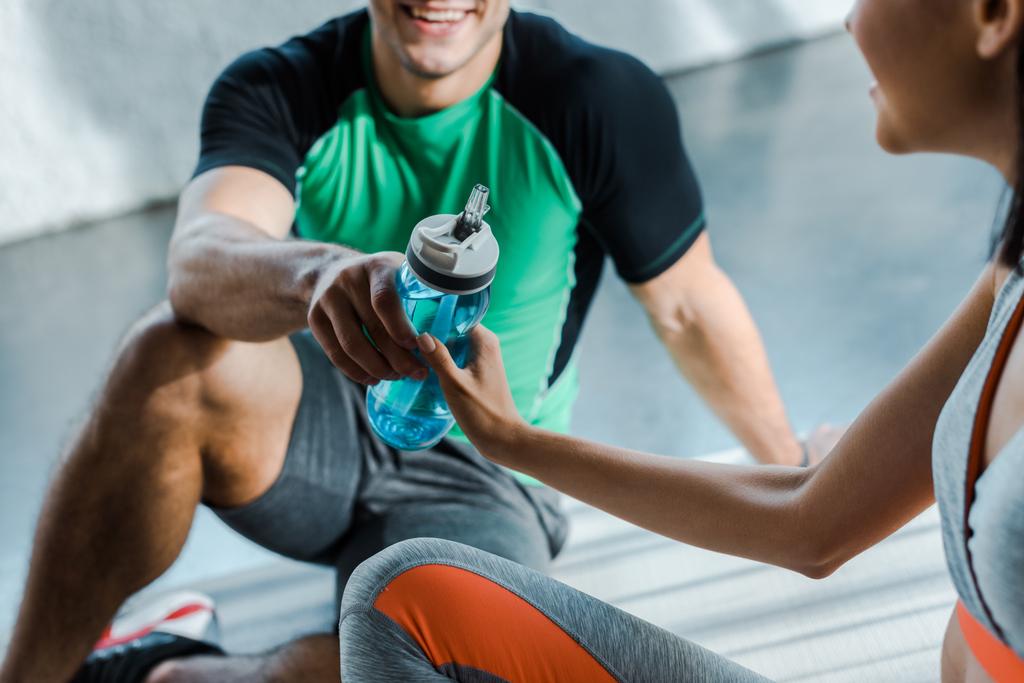 vue recadrée d'un sportif souriant donnant une bouteille de sport à une sportive dans un centre sportif
 - Photo, image