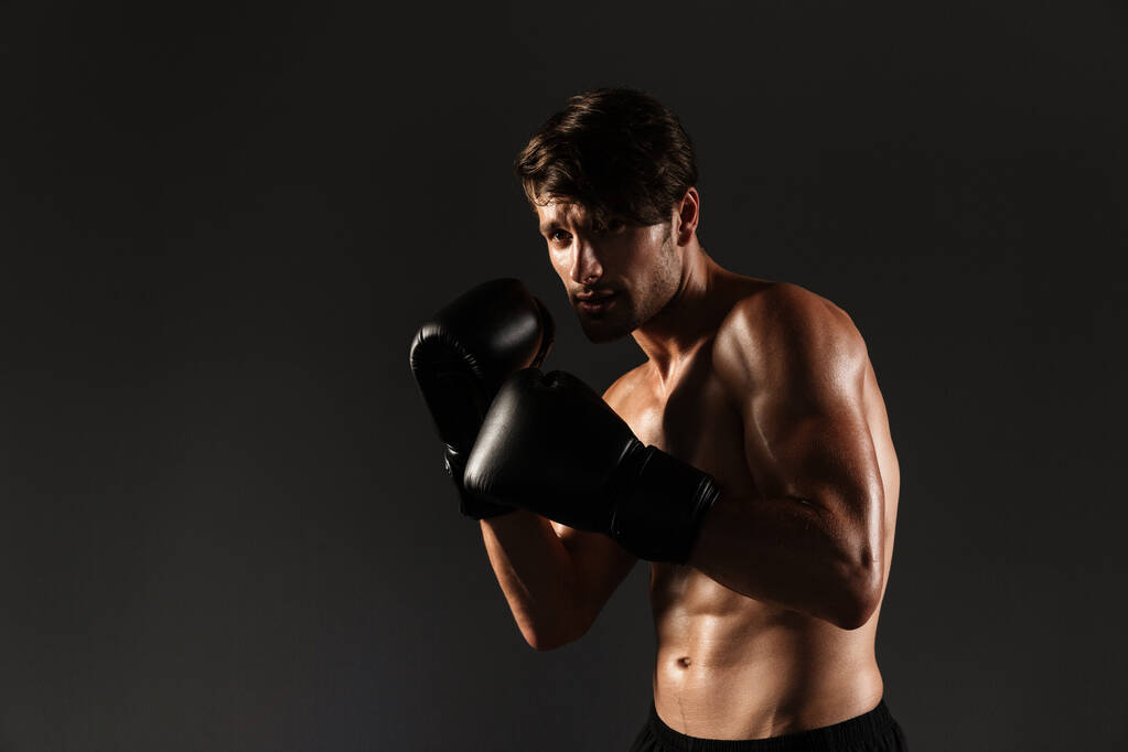 Guapo joven deportista fuerte boxeador en guantes hacer ejercicios de boxeo aislado sobre fondo negro de la pared
. - Foto, Imagen