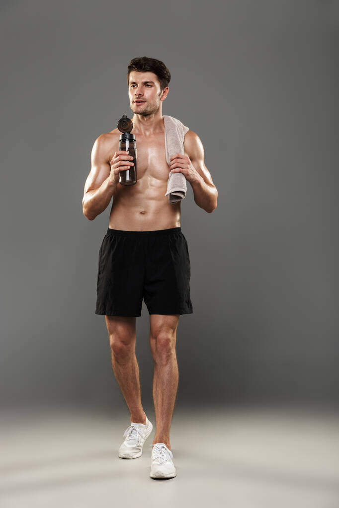 Handsome young strong sportsman isolated over grey wall background holding towel and bottle with water. - Photo, Image