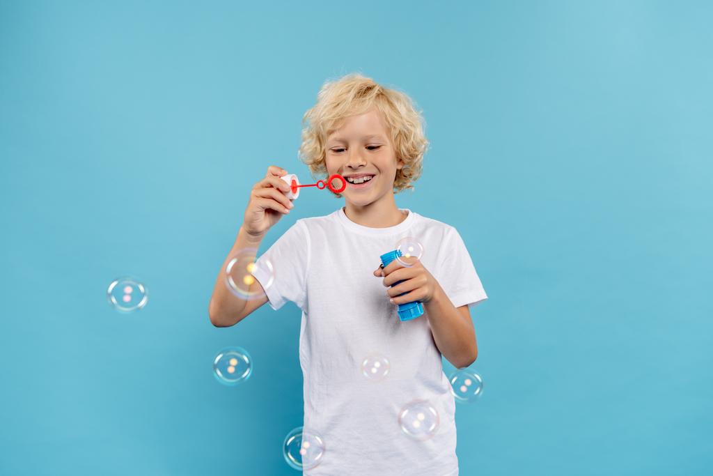 uśmiechnięty dzieciak w białej koszulce dmuchający bańki mydlane na niebieskim tle  - Zdjęcie, obraz