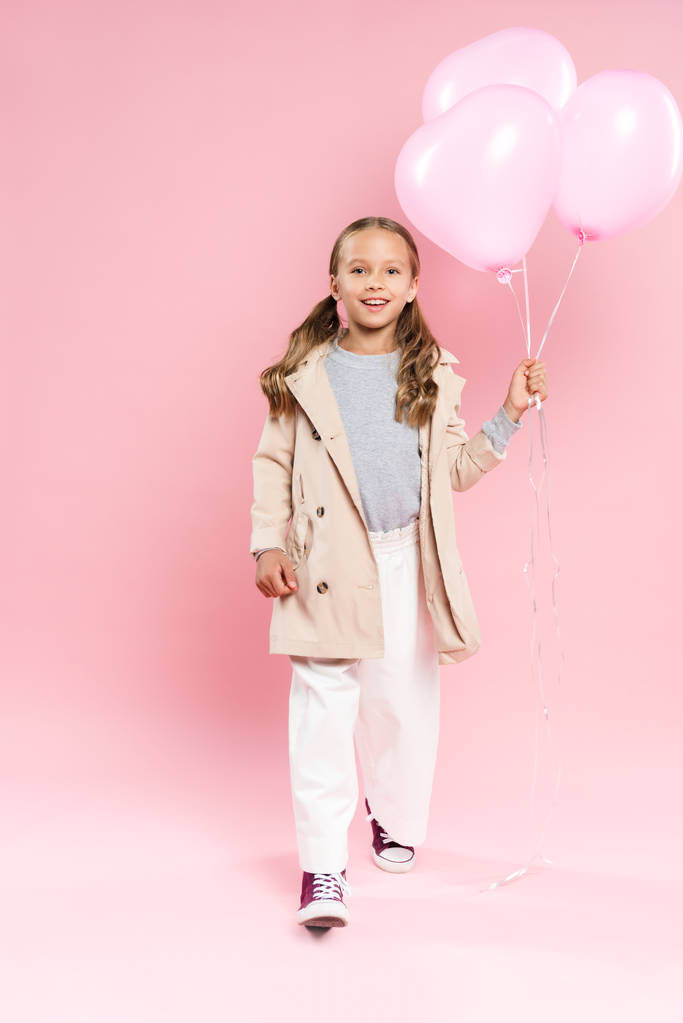 smiling kid in autumn outfit walking and holding balloons on pink background  - Photo, Image