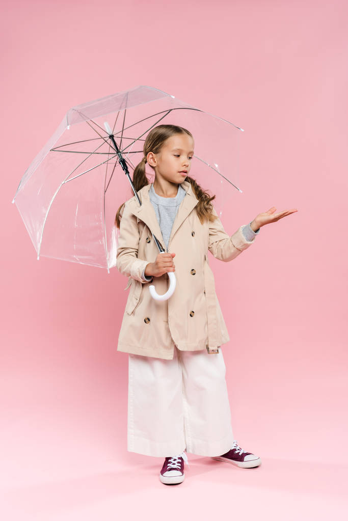 Kind im Herbst-Outfit mit ausgestreckter Hand, die Regenschirm auf rosa Hintergrund hält  - Foto, Bild