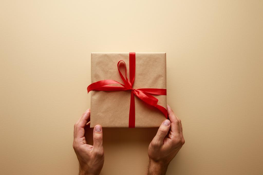 abgeschnittene Ansicht des Mannes mit Geschenkschachtel in Bastelpapier mit rotem Band auf beigem Hintergrund - Foto, Bild