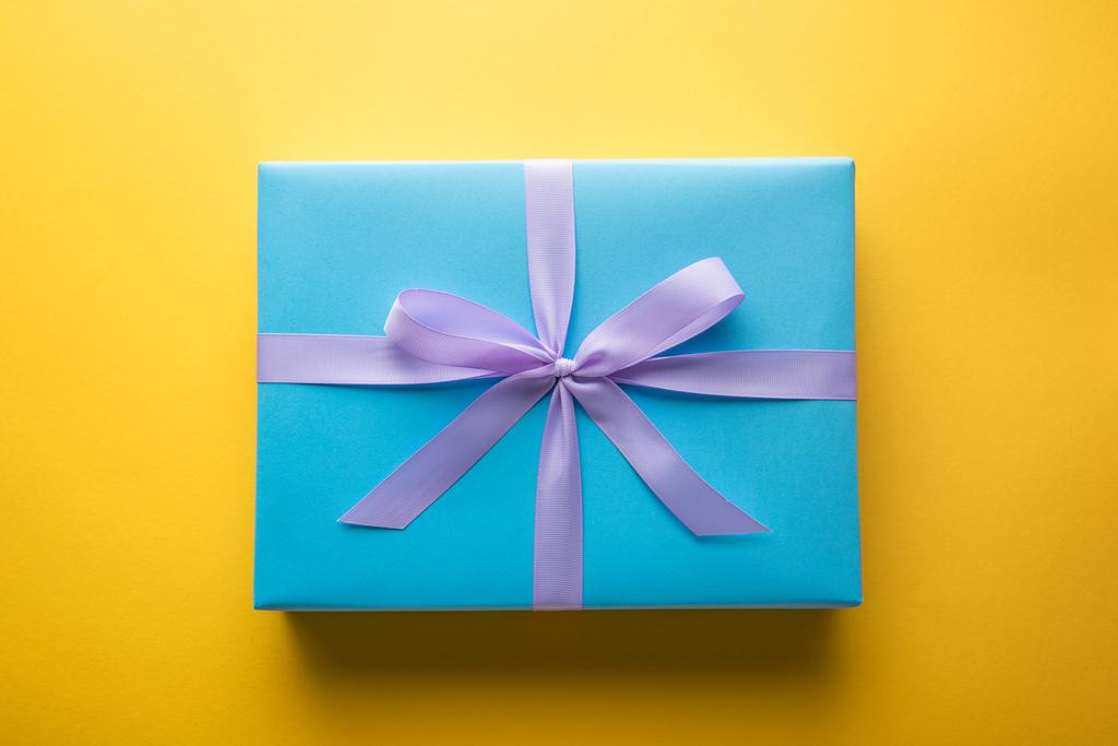 πάνω όψη του μπλε κουτί δώρου με βιολετί κορδέλα σε κίτρινο φόντο - Φωτογραφία, εικόνα