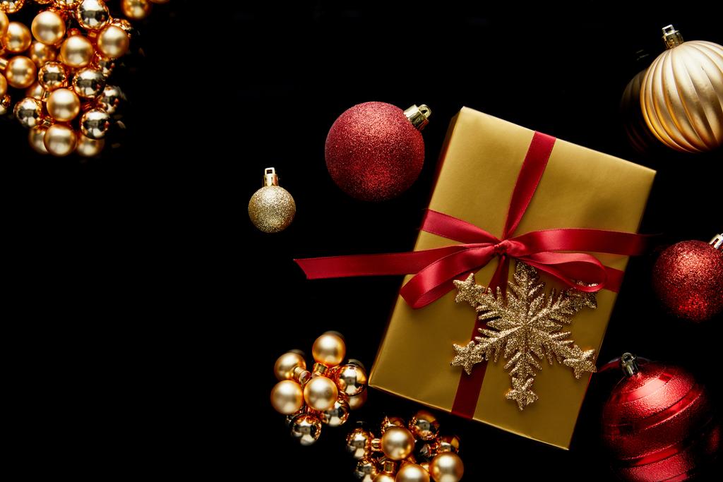 vrchní pohled na lesklý zlatý vánoční dárek s červenou stuhou a sněhovou vločkou v blízkosti ozdoby izolované na černé - Fotografie, Obrázek