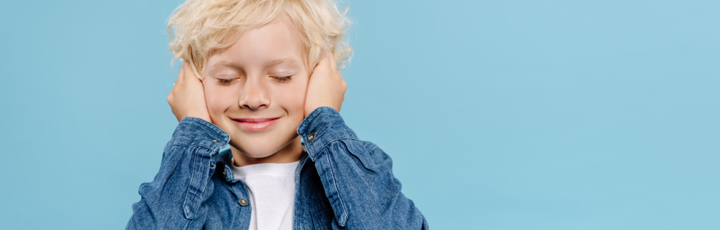 Panoramaaufnahme eines lächelnden und niedlichen Kindes mit geschlossenen Augen, das isoliert auf blauem Grund Ohren berührt  - Foto, Bild
