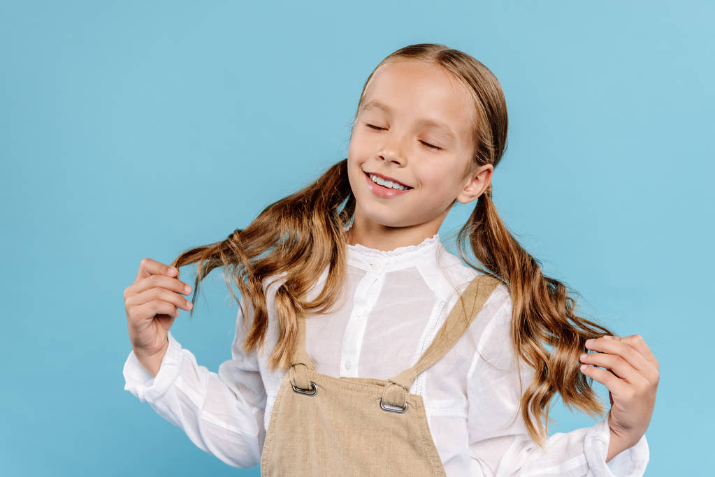 lächelndes und nettes Kind, das die Haare isoliert auf blau berührt  - Foto, Bild