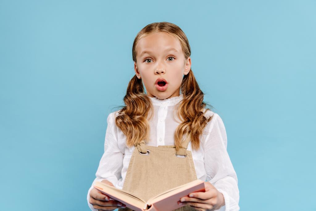 shocked and cute kid looking at camera and holding book isolated on blue  - Photo, Image