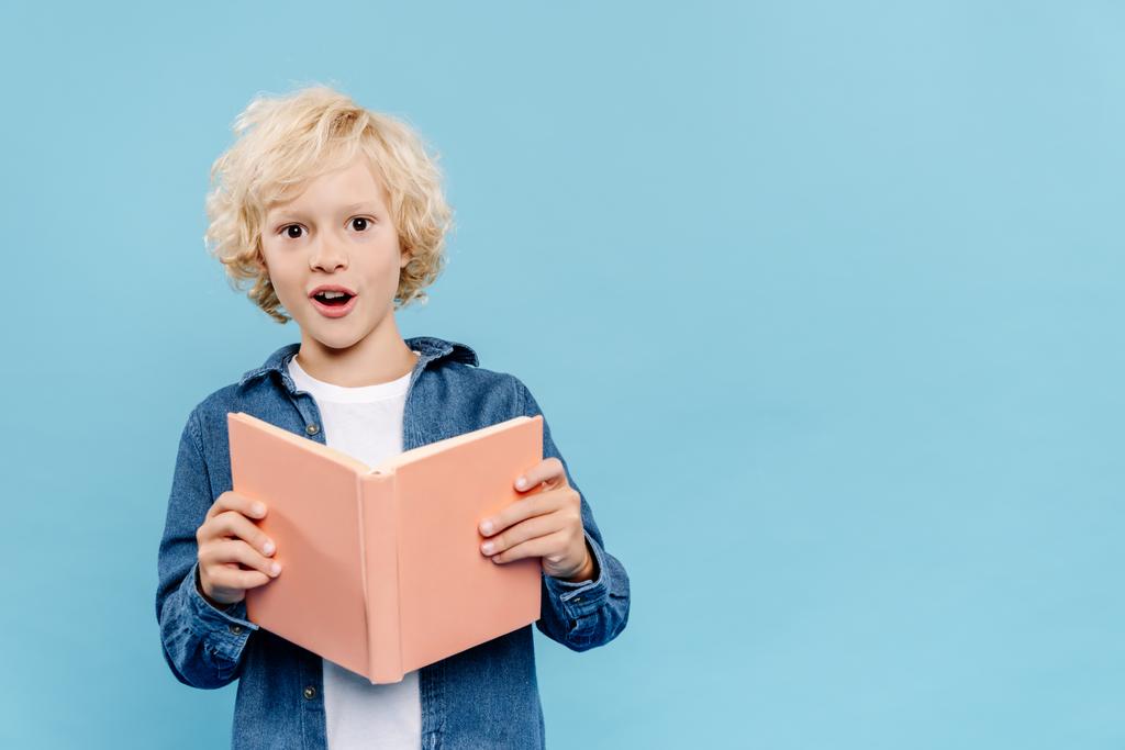 σοκαρισμένος και χαριτωμένο παιδί κρατώντας βιβλίο και κοιτάζοντας κάμερα απομονωμένη στο μπλε  - Φωτογραφία, εικόνα