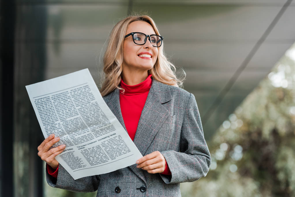 aantrekkelijke zakenvrouw in jas en bril glimlachen en houden krant  - Foto, afbeelding