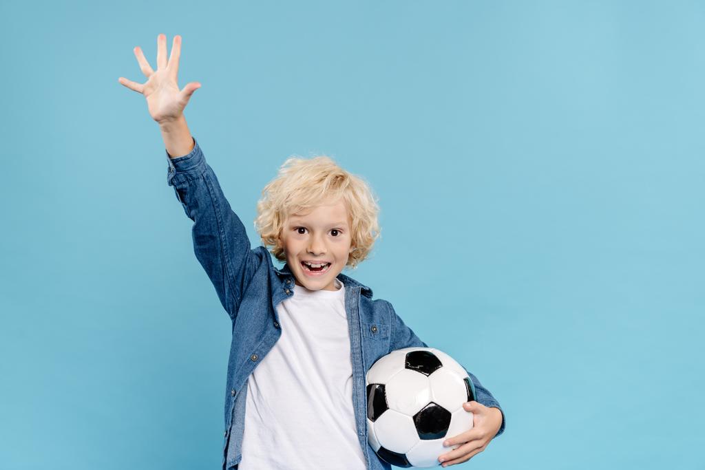 sorrindo e bonito garoto com mão estendida olhando para a câmera e segurando futebol isolado em azul
  - Foto, Imagem