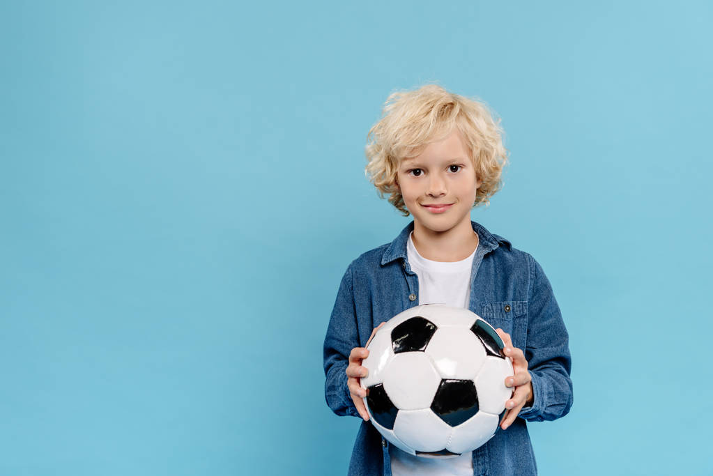 uśmiechnięty i słodki dzieciak patrząc w aparat i trzymając piłkę nożną odizolowany na niebiesko  - Zdjęcie, obraz