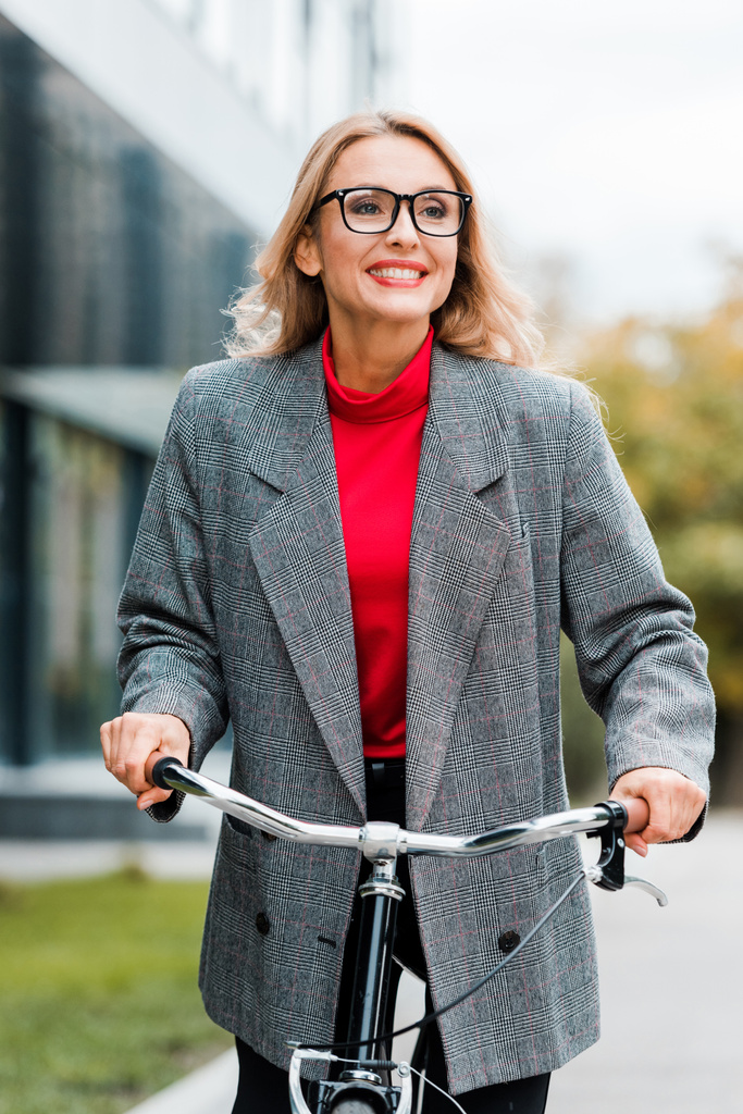 привлекательная деловая женщина в пальто и очках, улыбающаяся и катающаяся на велосипеде
  - Фото, изображение