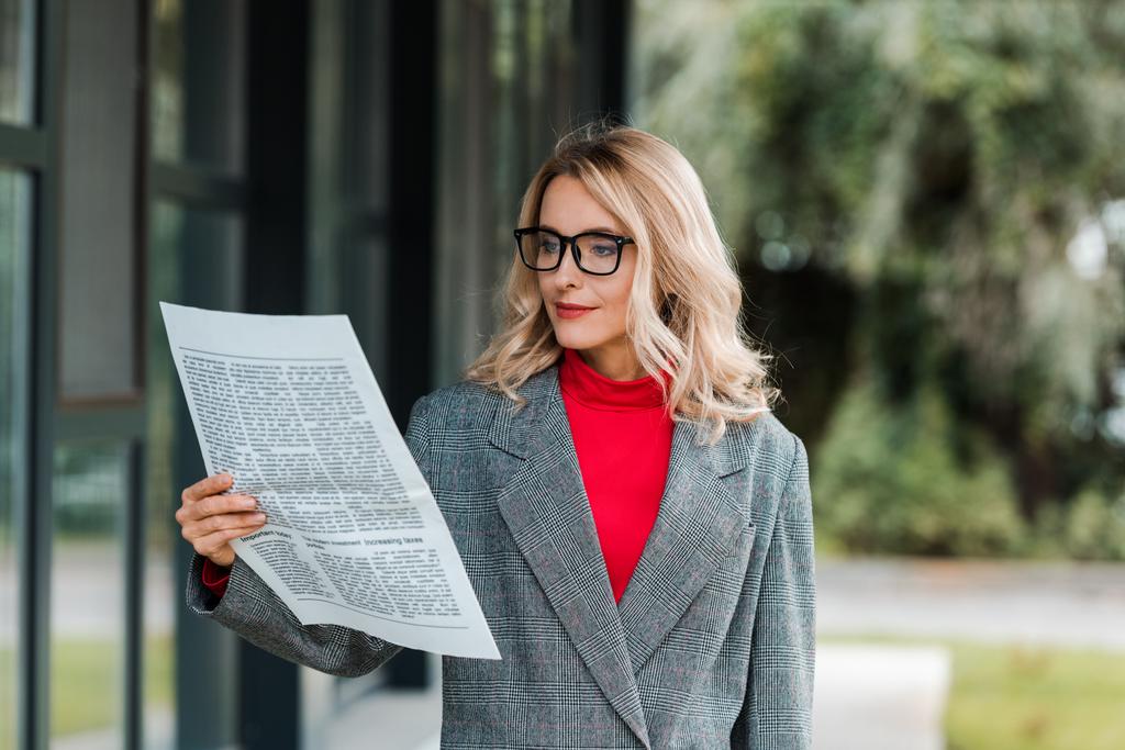 atrakcyjna bizneswoman w szarym płaszczu i okularach czytająca gazetę  - Zdjęcie, obraz