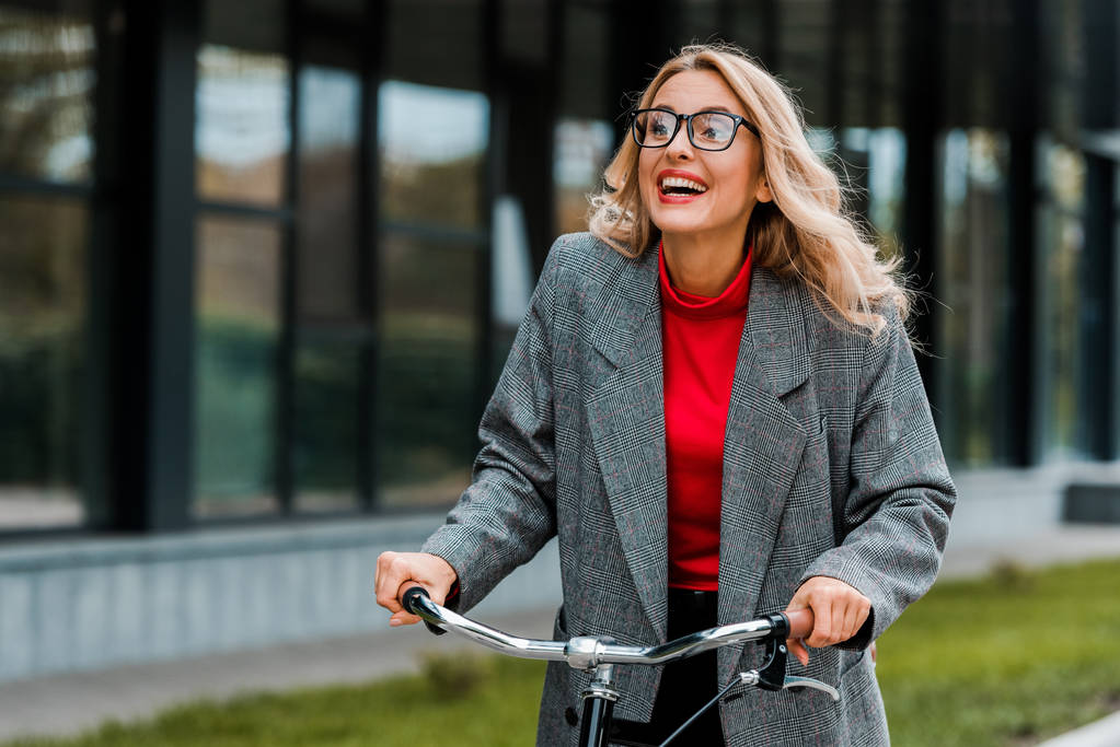 ελκυστική επιχειρηματίας σε παλτό και γυαλιά χαμογελώντας και ιππασία ποδήλατο  - Φωτογραφία, εικόνα