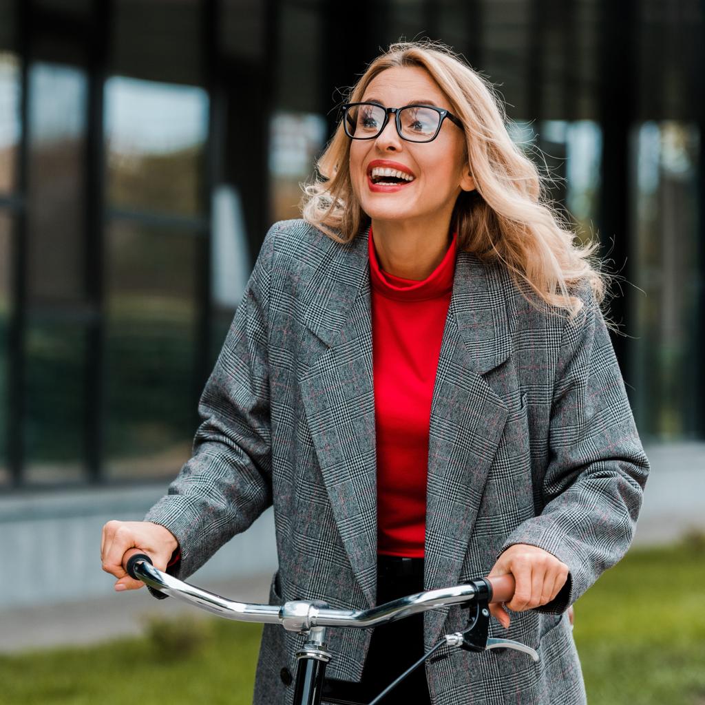 attractive businesswoman in coat and glasses smiling and riding bike  - Photo, Image
