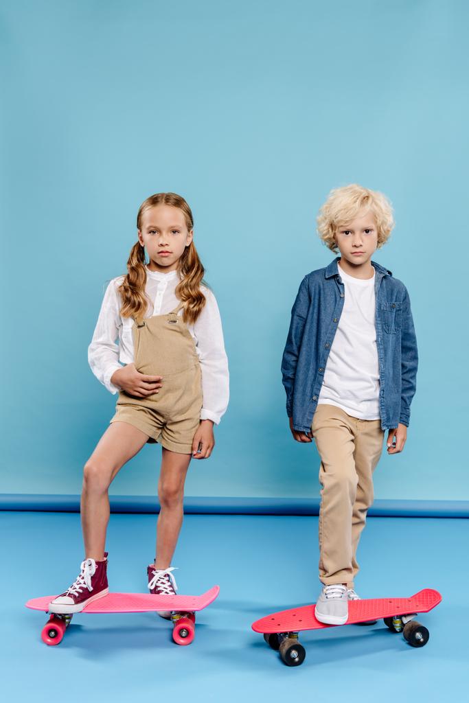 roztomilé děti při pohledu na kameru a stojí na penny desky na modrém pozadí  - Fotografie, Obrázek