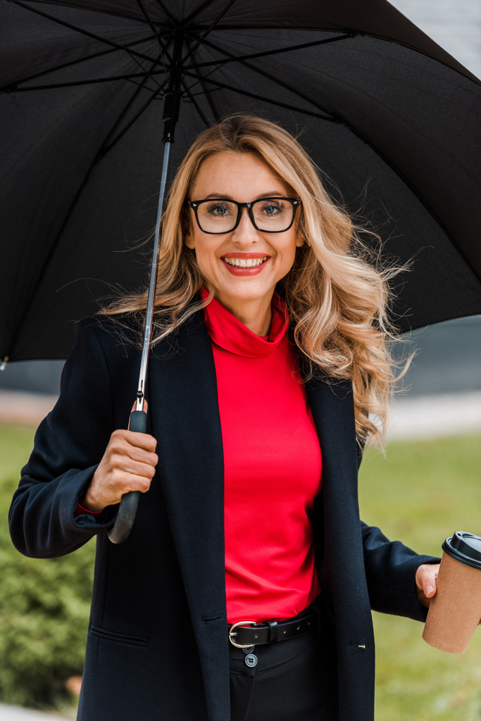 atrakcyjna i uśmiechnięta bizneswoman w płaszczu z parasolem i papierową filiżanką - Zdjęcie, obraz