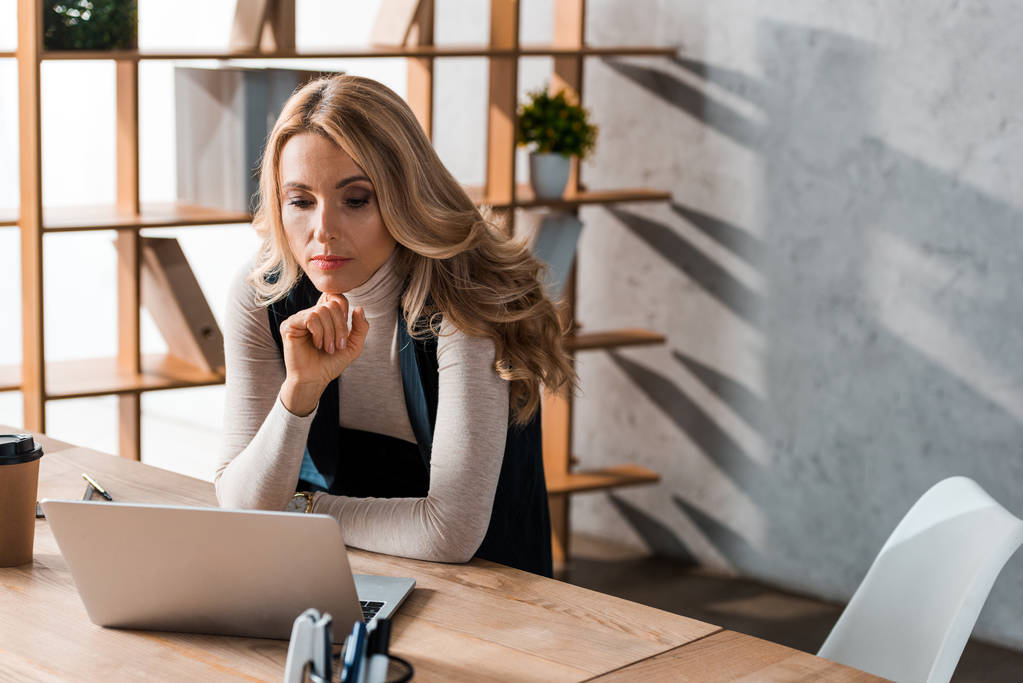 atrakcyjna i blondynka bizneswoman siedzi przy stole i patrzy na laptopa  - Zdjęcie, obraz