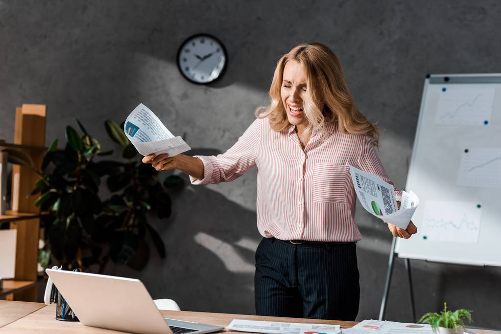 ελκυστική και θυμωμένη επιχειρηματίας με πουκάμισο κρατώντας χαρτιά και ουρλιάζοντας στο γραφείο  - Φωτογραφία, εικόνα