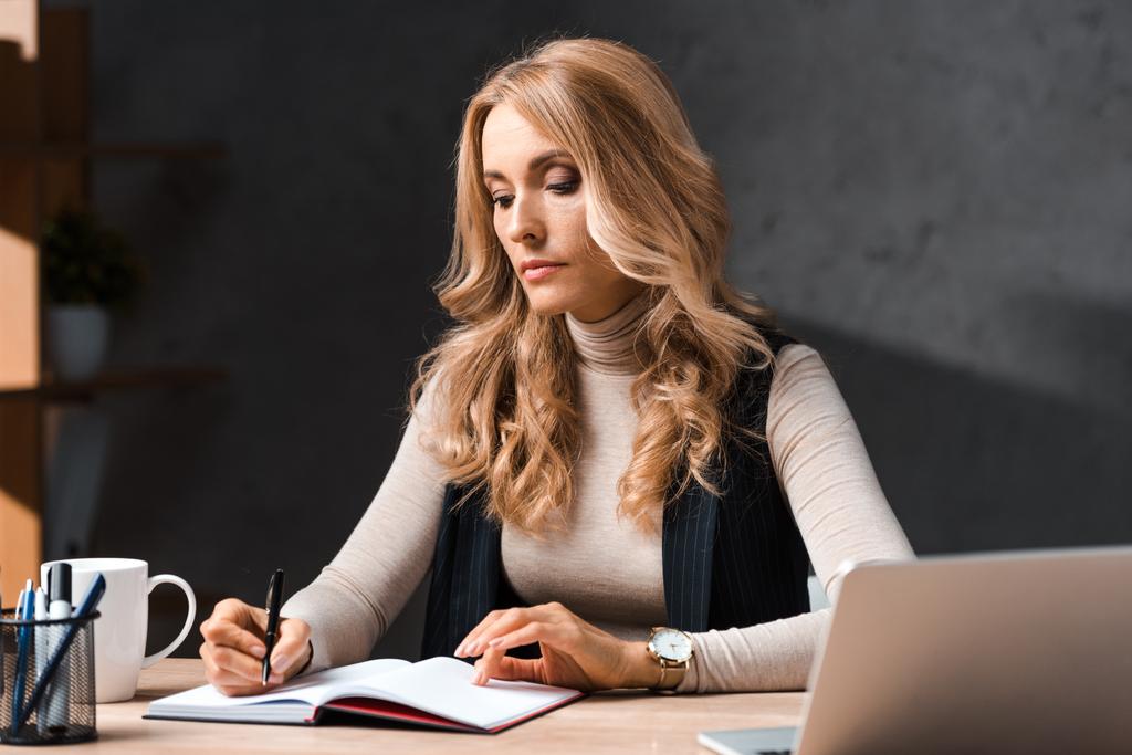 attraktive und blonde Geschäftsfrau sitzt am Tisch und schreibt in Notizbuch im Büro  - Foto, Bild