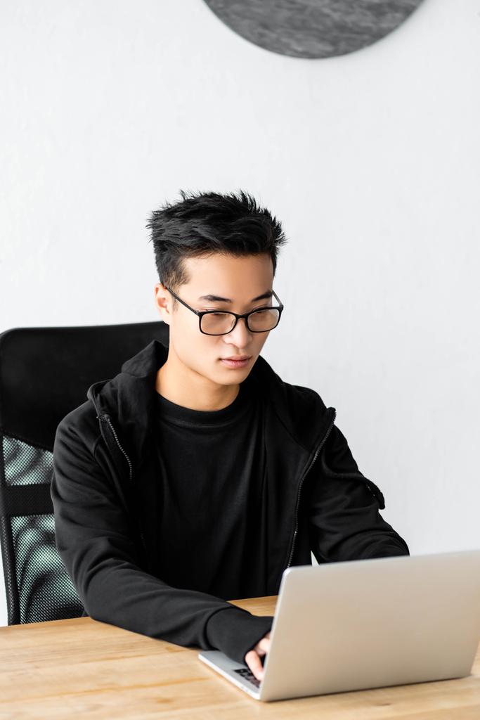 hacker asiático en gafas sentado en la mesa y el uso de ordenador portátil
  - Foto, imagen