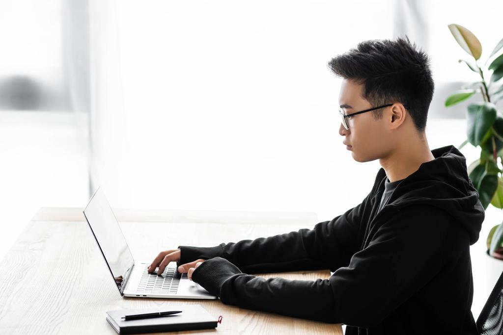 вид сбоку азиатского хакера в очках, сидящего за столом и использующего ноутбук
  - Фото, изображение
