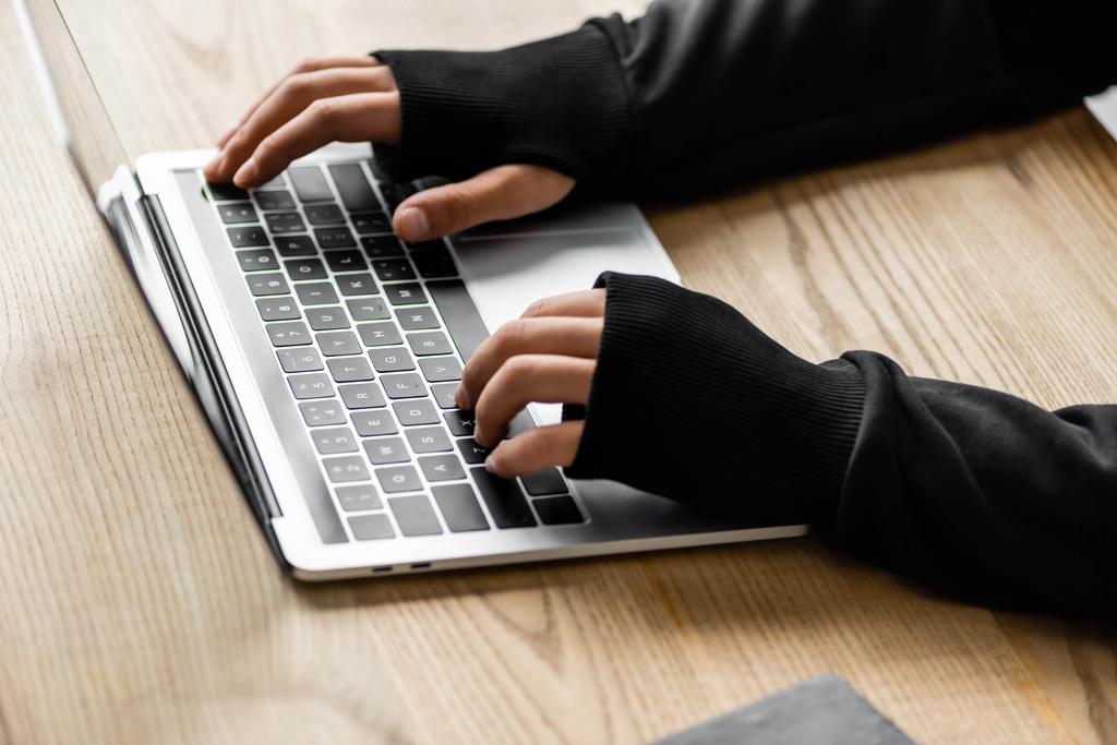 przycięty widok hakera siedzącego przy stole i korzystającego z laptopa  - Zdjęcie, obraz