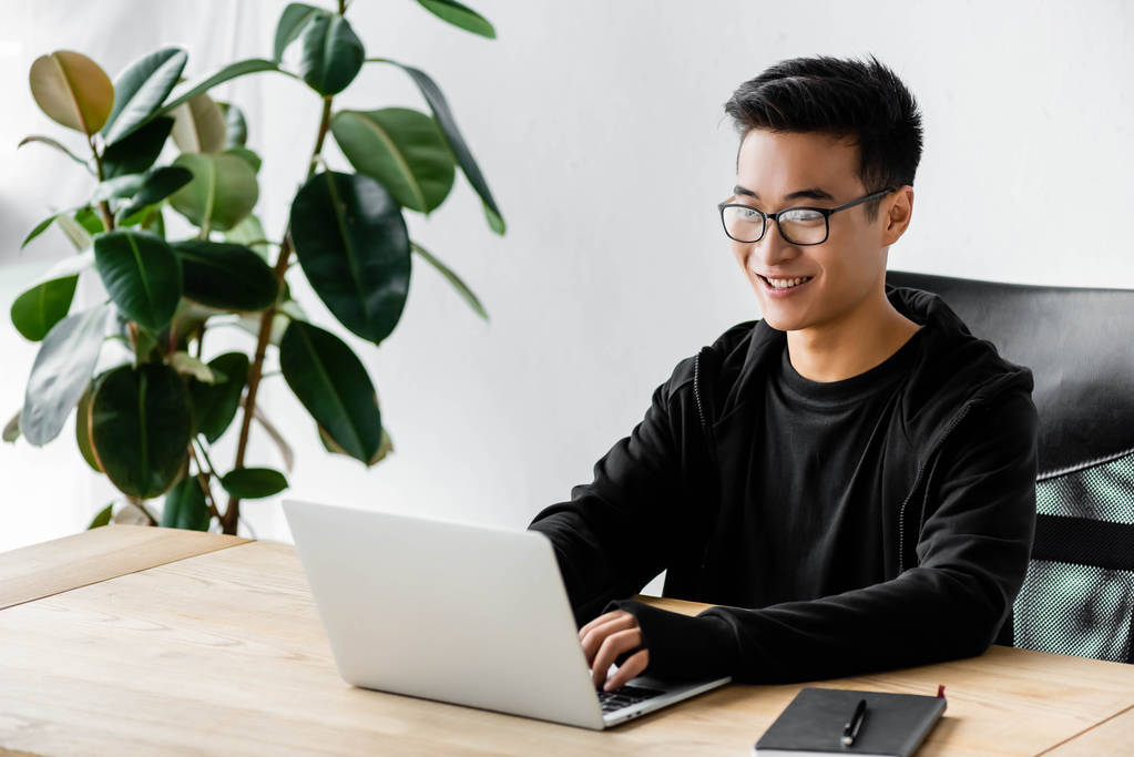 sorrindo asiático hacker no óculos sentado no mesa e usando laptop
  - Foto, Imagem
